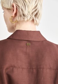 Hnedá látková blúzka s golierom, so zlatým logom na zadnej strane a viditeľnými švami. Nosia sa veľké kruhové náušnice.