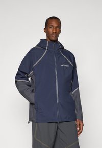Navy and gray waterproof jacket with a zip front, hood, and reflective accents. Features a lightweight, smooth texture and has a relaxed fit.
