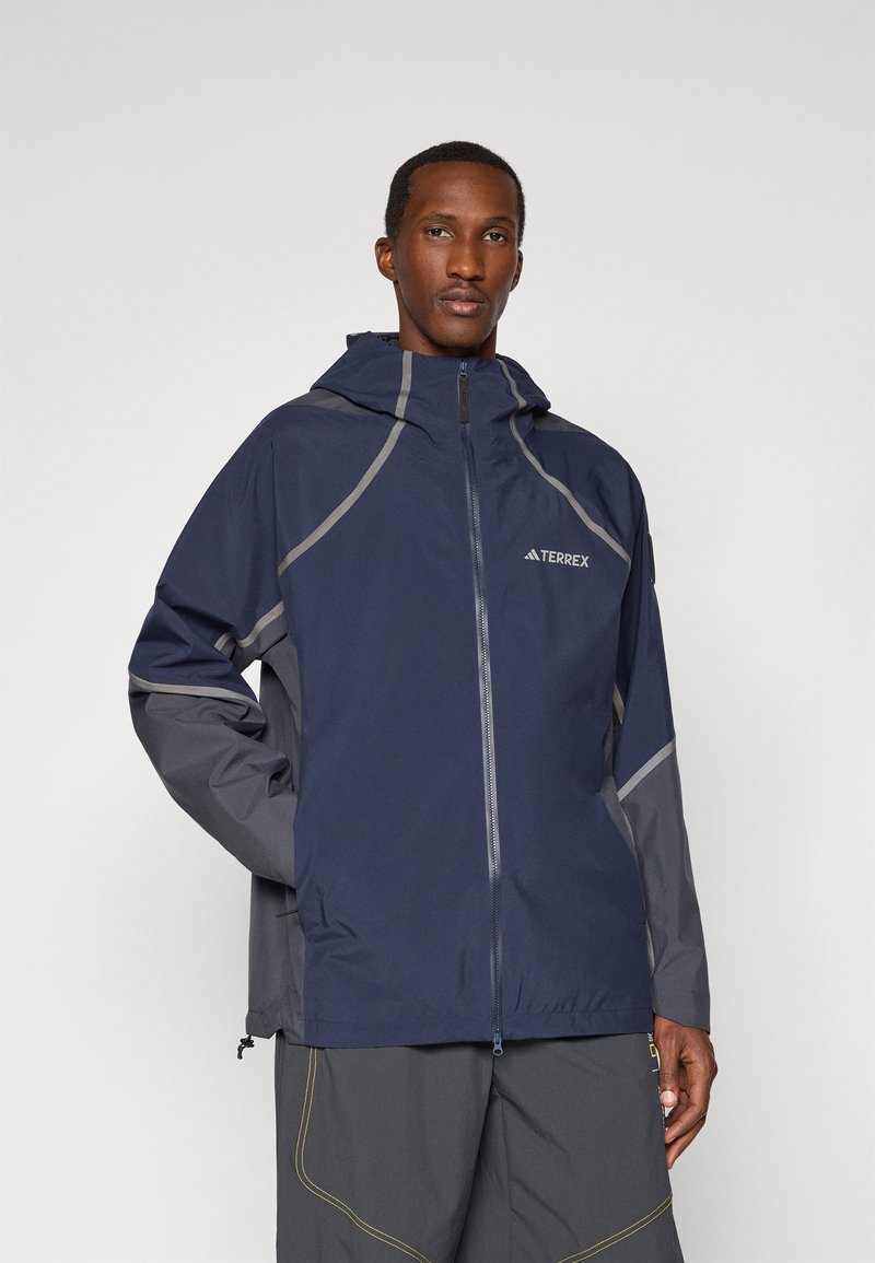 Navy and gray waterproof jacket with a zip front, hood, and reflective accents. Features a lightweight, smooth texture and has a relaxed fit.