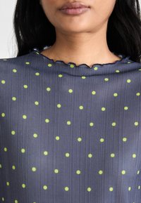 Top trasparente blu navy con texture a costine, caratterizzato da grandi pois verdi e un colletto scallop, dettagli visibili sul tessuto e sul motivo.