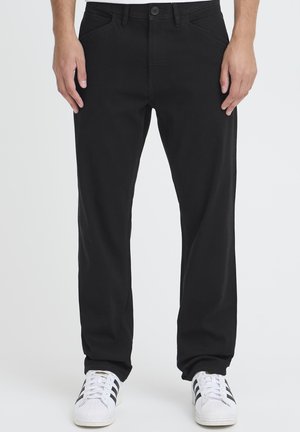 Pantalon noir en coton, coupe droite, poches avant, fermeture à boutons, porté avec des baskets blanches.