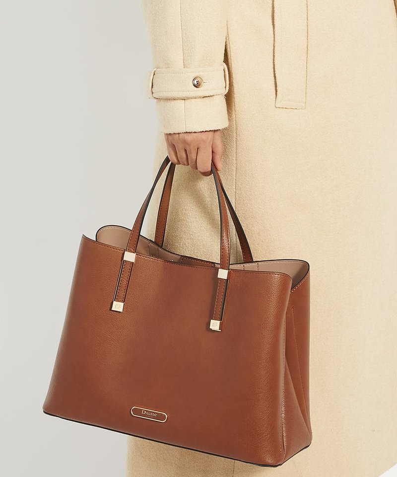 Main tenant un grand sac cabas en cuir marron avec double poignées, porté sur un manteau long beige avec poignets boutonnés.