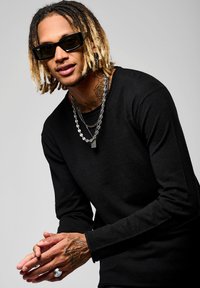 Camicia nera a maniche lunghe con una finitura testurizzata, catene d'argento sovrapposte, occhiali da sole e tatuaggi visibili su mani e collo.