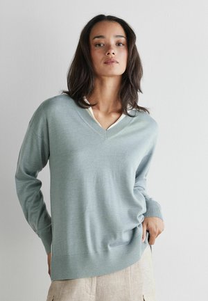 Femme aux longs cheveux foncés portant un pull bleu clair à col en V et un pantalon beige, debout contre un mur blanc uni.