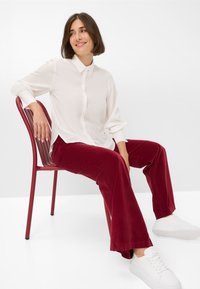 Witte button-up shirt met opgerolde mouwen, gecombineerd met rode flared corduroy broek. Persoon zittend op een rode stoel, draagt witte sneakers.
