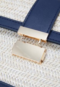 Cinturino in pelle blu navy con una fibbia rettangolare dorata su uno sfondo di paglia intrecciata e testurizzata. Superfici lisce contrastano con la trama intrecciata.