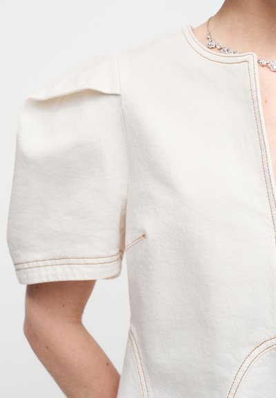 Εκρού ένδυμα με κοντά φουσκωτά μανίκια, διακοσμημένο με βαθύ V ντεκολτέ και αντίθετη ραφή. Ομαλή υφή βαμβακιού με ελάχιστες λεπτομέρειες.