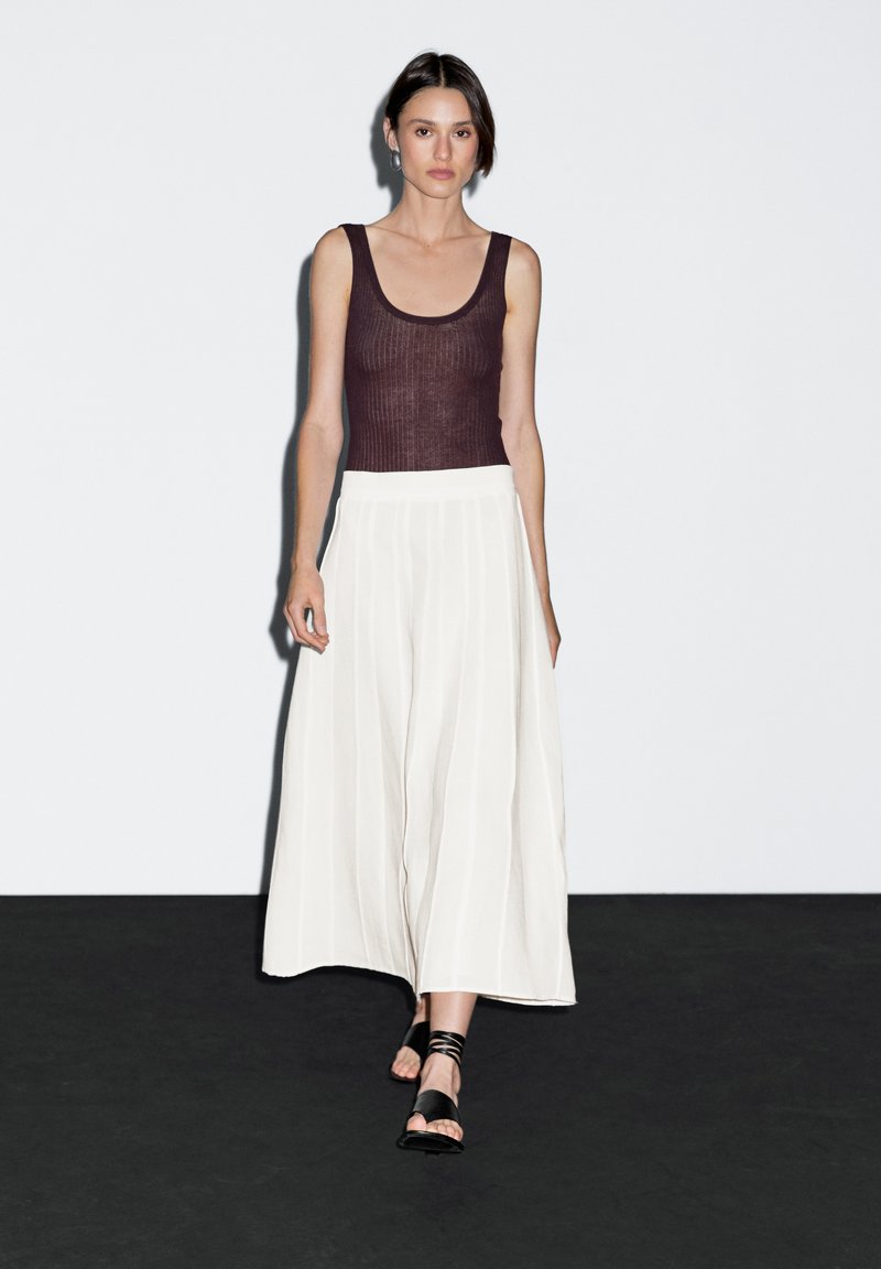 Una modella cammina con sicurezza indossando un top aderente marrone e culottes bianche fluide, sullo sfondo minimalista bianco e nero.