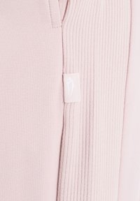 Hellrosa Stoff mit glatter und gerippter Struktur, verbunden durch eine Naht, mit einem kleinen weißen Etikett mit dezentem Logo.