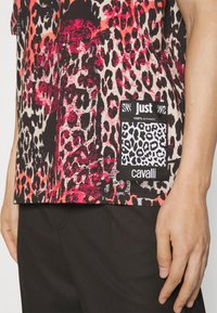 Leopardmönstrad skjorta med röda och svarta detaljer, med en "Just Cavalli 100% äkta" etikett vid fållen, bärs med svarta byxor.