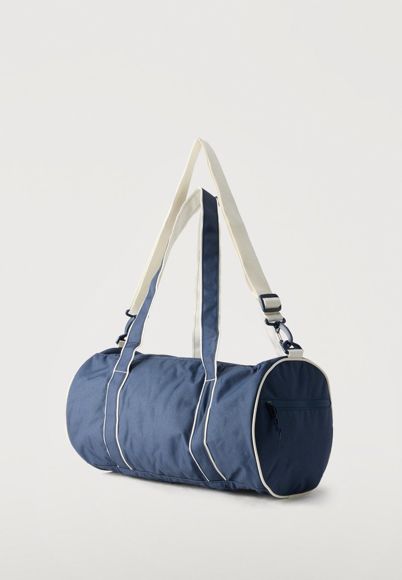 Marineblauer zylindrischer Sportbeutel mit weißem Besatz, doppelten Griffen, verstellbarem Schulterriemen und einem Reißverschlussfach an einem Ende.