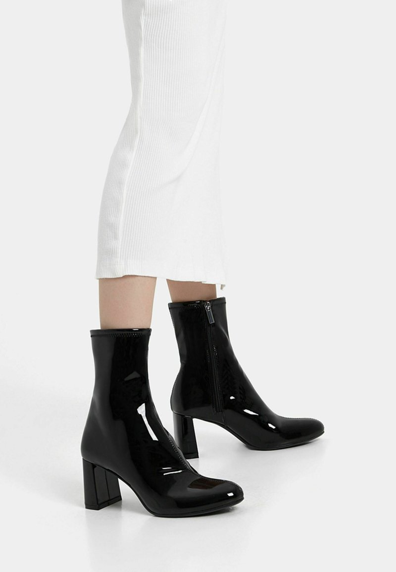 Černé lakované kotníkové boty s blokovým podpatkem, se zipem na boku, hladkým povrchem a zaoblenou špičkou. Nosené s bílými žebrovanými šaty.