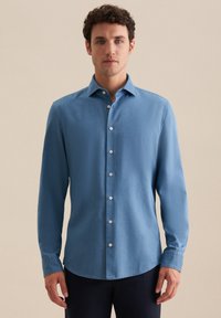 Uomo in piedi che indossa una camicia a maniche lunghe azzurro chiaro e pantaloni scuri, contro uno sfondo beige semplice.