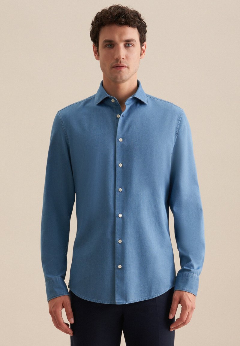 Uomo in piedi che indossa una camicia a maniche lunghe azzurro chiaro e pantaloni scuri, contro uno sfondo beige semplice.