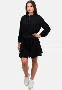 Abito nera con bottoni, maniche lunghe e cintura in vita, realizzato in un tessuto texturizzato e con gonna svasata, indossato con sneakers bianche.