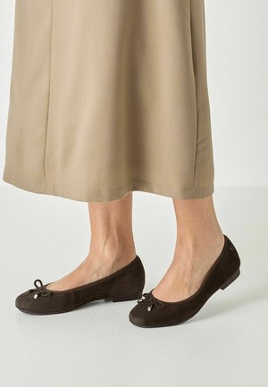 Kvinde iført mørkebrune ruskind ballet flats med små sløjfedetaljer, stående på lyst gulv ved siden af en beige midi nedsmøget nederdel.
