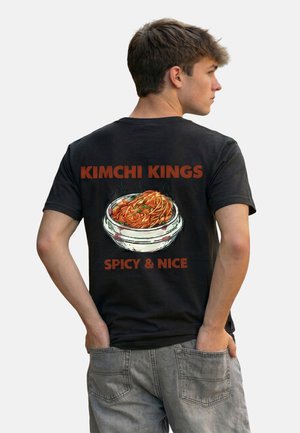 Junger Mann trägt ein schwarzes T-Shirt mit einer dampfenden Schüssel Kimchi-Nudeln und dem Text „KIMCHI KINGS SPICY & NICE“ auf dem Rücken.
