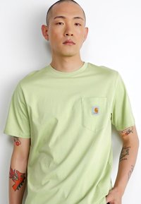 T-shirt en coton vert clair avec un col rond, des manches courtes et une poche poitrine arborant une étiquette logo. Texture lisse, design décontracté.