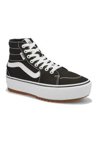 Zapatilla alta de lona negra con cordones blancos y logo, que cuenta con una gruesa suela blanca de plataforma y una suela de goma marrón con un patrón de tracción.