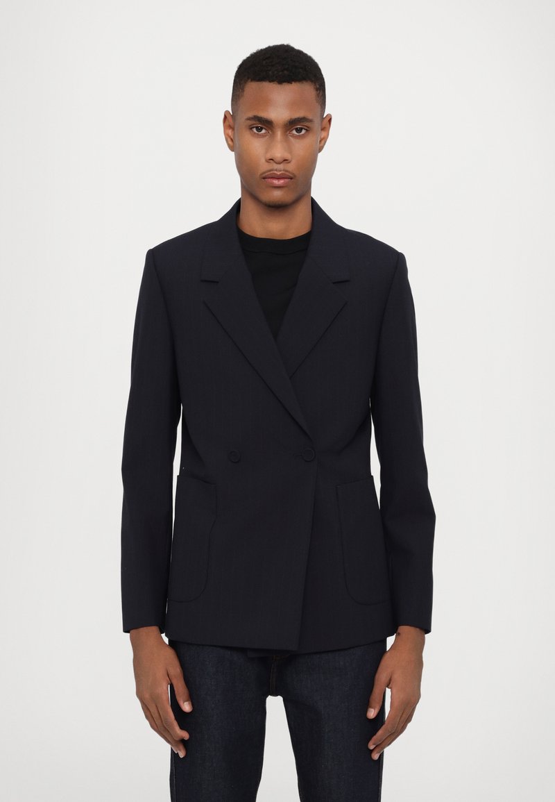Marineblaues, doppelt geknöpftes Blazer mit Reverskragen, zwei Vorderaschen und einer strukturierten Passform, getragen über einem schwarzen Hemd und dunklen Jeans.