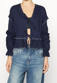 Blusa cropped di colore blu navy con orlo e polsini scallop, caratterizzata da lacci frontali e dettagli azzurri. Tessuto morbido con una vestibilità rilassata.