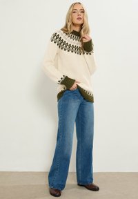Cremiger Pullover mit grünem gemustertem Einsatz und gerippten Bündchen, kombiniert mit blauen weit geschnittenen Jeans und braunen Stiefeln, vor einem neutralen Hintergrund.