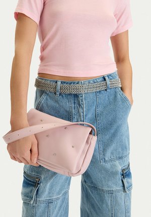 Persona que lleva una camiseta rosa claro recortada y vaqueros cargo azules con cinturón de cadena plateada, sosteniendo un pequeño bolso rosa con tachuelas.