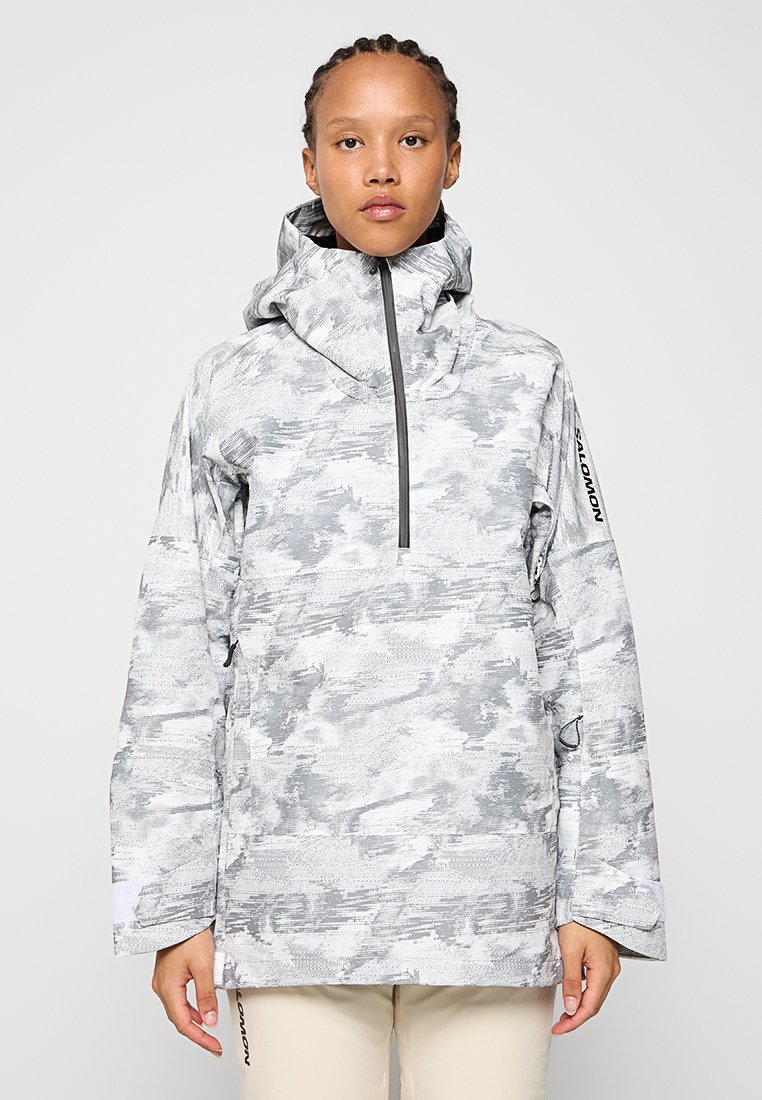 Chaqueta impermeable gris y blanca con un patrón, cuello alto, cierre de cremallera y mangas largas. Cuenta con capucha y un discreto detalle de logo.