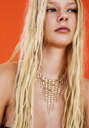 Giovane donna con lunghi capelli biondi che indossa una collana a catena dorata a strati su uno sfondo arancione.