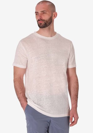 Uomo con la barba che indossa una maglietta beige chiaro a maniche corte e pantaloni blu, in piedi con una mano in tasca, guarda di lato.