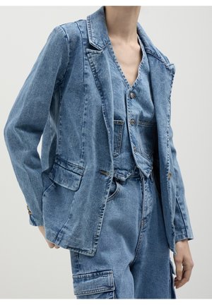 Persoon die een lichtblauwe denim blazer draagt, een dichtgeknoopte denim bodywarmer en hooggetailleerde denim cargobroek met zakken, tegen een effen achtergrond.