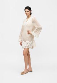 Γυναίκα που φοράει ένα μπεζ πλεκτό φόρεμα με φλοράλ δαντελένιο τελείωμα, στέκεται με το ένα χέρι στη μέση της, φορώντας μπεζ επίπεδα σανδάλια σε λευκό φόντο.