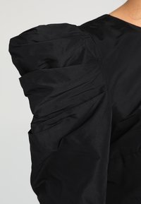 Černá blůza s strukturovaným nabíraným rukávem a hladkou texturou. Materiál vypadá lehký a matný.
