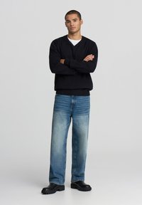 Uomo in piedi con le braccia incrociate, che indossa un maglione nero, jeans blu sbiaditi e stivali neri massicci, su uno sfondo neutro.