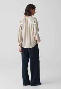 Beige blouse met lange mouwen met een geplooid detail op de rug, gecombineerd met een losvallende donkerblauwe broek. Zachte stof met een gladde textuur.