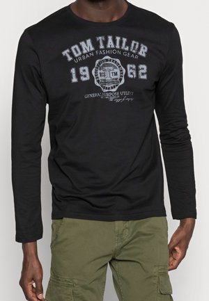 Hombre con camiseta negra de manga larga con gráfico "Tom Tailor 1962" y pantalones cargo verdes oliva, posando frente a un fondo liso.