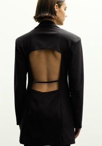 Schwarze taillierte Blazerjacke mit langen Ärmeln und einem offenen Rückendesign, das mit einem geschwungenen Ausschnitt und einem gebundenen Detail in der Taille versehen ist. Glatte Textur.