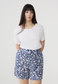 T-shirt bianca con dettagli in pizzo sulle maniche, abbinata a pantaloni corti blu con motivo floreale. Il design presenta accenti bianchi su uno sfondo scuro.