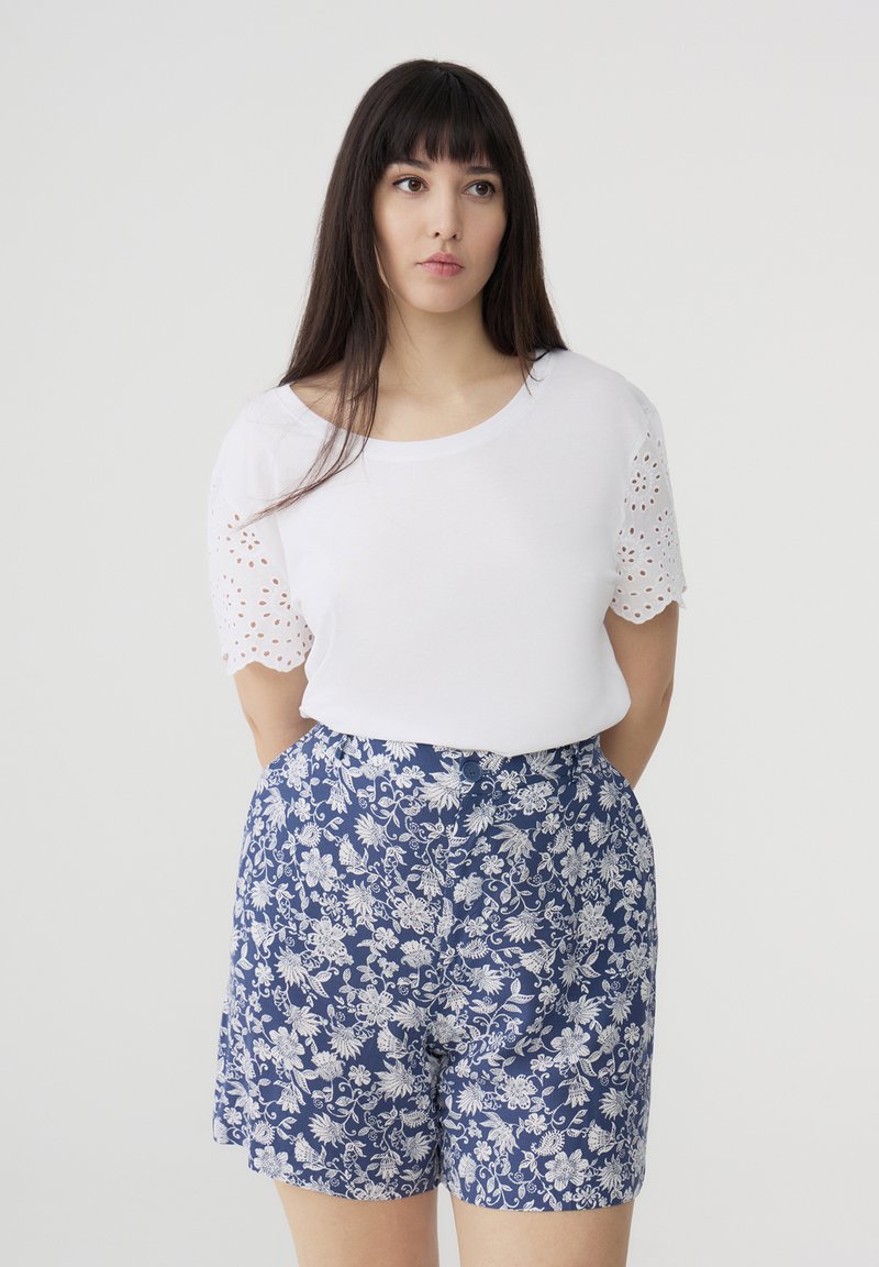 T-shirt bianca con dettagli in pizzo sulle maniche, abbinata a pantaloni corti blu con motivo floreale. Il design presenta accenti bianchi su uno sfondo scuro.