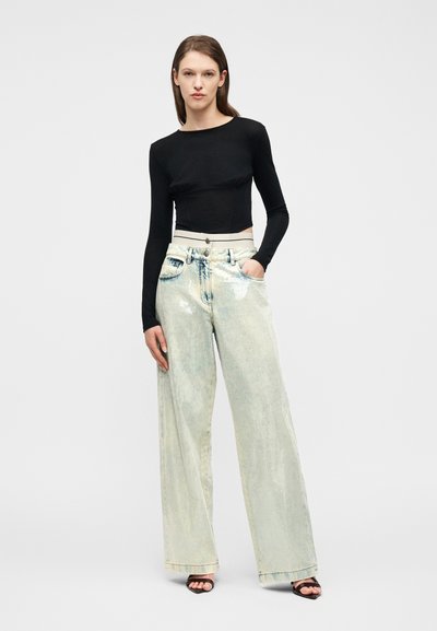 Γυναίκα που στέκεται, φοράει εφαρμοστό μαύρο μακρυμάνικο τοπ, ανοιχτόχρωμα τζιν με φαρδιά μπατζάκια και ψηλόμεση μέση, καθώς και μαύρα πέδιλα με τακούνι και ανοιχτά δάχτυλα, μπροστά σε απλό φόντο.