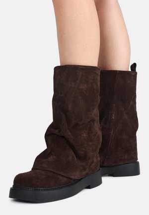 Bottes mi-mollet en daim brun foncé doux avec semelles en caoutchouc noires plates, portées sur des jambes nues sur un fond blanc uni.