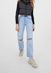 Woman wearing light blue ripped jeans, cream chunky lace-up boots, black long-sleeve top, and black padded cropped vest against white background.