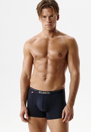 Męski muskularny mężczyzna z krótkimi brązowymi włosami, ubrany w czarne bokserki Atlantic, stojący na gładkim białym tle.