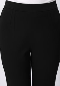 Μαύρο παντελόνι με ψηλή μέση κατασκευασμένο από ομαλό, ελαστικό υλικό, με δύο πτυχώσεις στο μπροστινό μέρος και απλή, καθαρή σχεδίαση. Χωρίς εμφανή μεταλλικά στοιχεία.