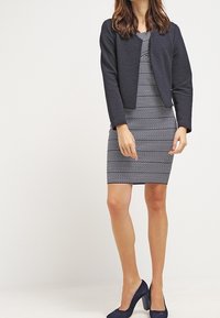 Námořnická texturovaná zkrácená bunda přes přiléhavé šaty se zigzagovým vzorem. Tmavě modré vysoké podpatky doplňují celý outfit. Hladká látka a dokonale padnoucí střih.
