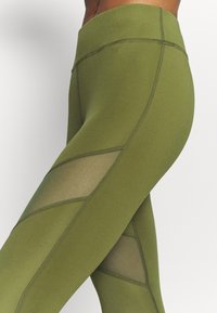 Leggings verde oliva con pannelli in rete e dettagli delle cuciture a contrasto. Tessuto morbido e elastico con una silhouette aderente.
