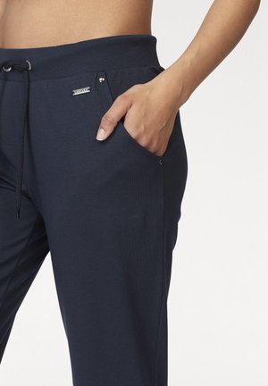 Person’s hand resting in the front pocket of navy blue drawstring pants with a metal logo tag on the waistband.