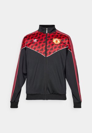 Čierna bunda na zips s červeným geometrickým vzorom, bielymi akcentmi a logom Manchester United. Má vysoký golier a rukávy so tromi pruhmi.