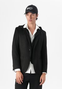 Schwarze, strukturierte Jacke mit Kapuze, zwei Fronttaschen und Knopfverschluss. Getragen über einem weißen Hemd und kombiniert mit einer schwarzen Mütze.