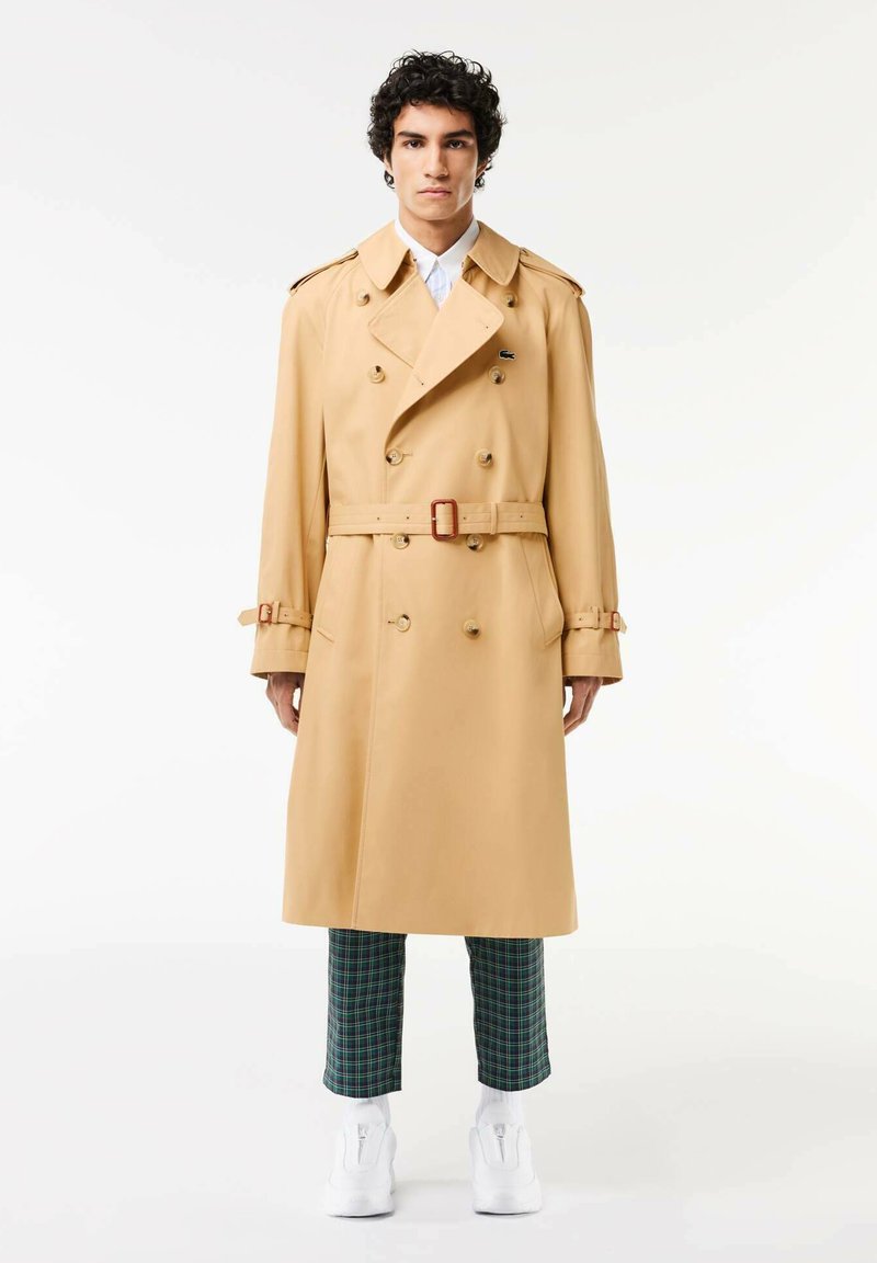 Manteau beige à double boutonnage avec de gros boutons, épaulettes et ceinture à la taille. Le mannequin porte un pantalon à carreaux verts et des chaussures blanches.